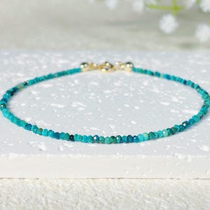 May include: A delicate bracelet featuring small, faceted turquoise beads strung together. The beads are a vibrant teal color, with hints of darker blue and green. The bracelet has a gold-colored clasp.