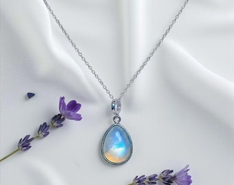 May include: A silver necklace with a teardrop-shaped pendant. The pendant features a moonstone with blue, white, and yellow hues. Lavender sprigs are placed near the necklace on a white fabric background.