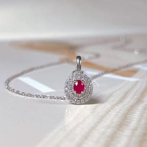 May include: A silver pendant necklace featuring a teardrop-shaped pendant. The pendant has a vibrant red oval gemstone at its center, surrounded by a halo of smaller, sparkling white gemstones. The necklace chain is delicate and silver.
