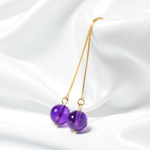 May include: A pair of dangle earrings featuring two round, deep purple amethyst beads. The beads are suspended from delicate gold chains, creating a minimalist and elegant design. The earrings are set against a white, silky background.