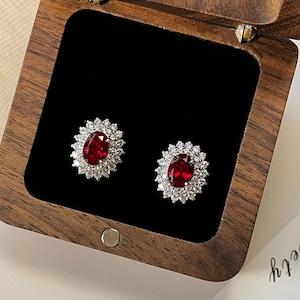 May include: A pair of ruby and diamond stud earrings displayed in a wooden jewelry box. Each earring features a central oval ruby surrounded by a halo of smaller, clear diamonds. The box is open, revealing a black velvet interior.