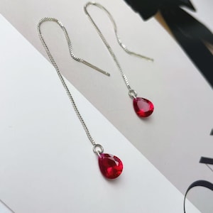 May include: A pair of silver drop earrings with a chain design. Each earring features a vibrant, teardrop-shaped, red gemstone. The earrings are displayed on a white surface with a black bow in the background.
