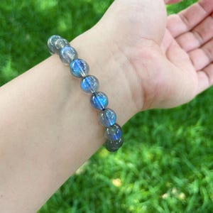 May include: A beaded bracelet featuring iridescent blue and gray beads. The bracelet is made of round, polished stones, and is worn on a wrist against a backdrop of green grass.