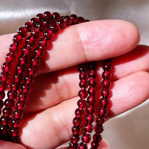 May include: A strand of deep red garnet beads, strung together in a long necklace. The beads are smooth and polished, with a slight shine.