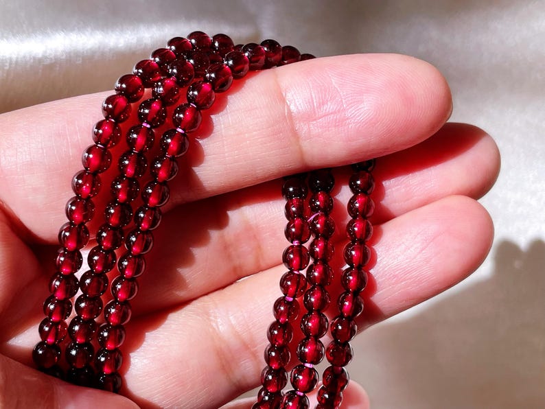 May include: A strand of deep red garnet beads, strung together in a long necklace. The beads are smooth and polished, with a slight shine.