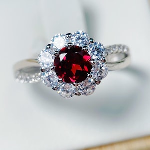 May include: A silver ring featuring a large, round, deep red gemstone surrounded by a halo of smaller, clear gemstones. The ring's band is also adorned with small, clear gemstones, creating a sparkling, elegant design.