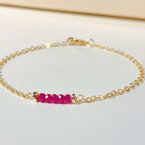 May include: Delicate gold chain bracelet featuring a row of five vibrant, faceted ruby gemstones. The bracelet has a simple, elegant design with a lobster clasp closure. The gold chain is thin and delicate.
