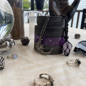 Può includere: Una collezione di oggetti mistici tra cui una collana con ciondolo a forma di luna e sole, una collana con ciondolo di cristallo, anelli, un cubo decorativo e occhiali da sole. Gli oggetti sono disposti su una tovaglia bianca.