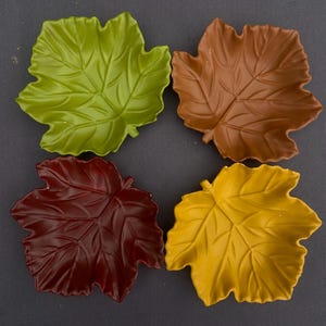 Op de afbeelding: Vier blad-vormige schalen in groen, bruin, bordeauxrood en geel. De schalen hebben gedetailleerde aderpatronen en zijn gerangschikt op een grijze ondergrond. Deze decoratieve schalen zijn ideaal voor het serveren van snacks of als herfstdecoratie.