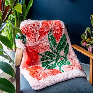 Könnte beinhalten: Eine rosa und orangefarbene Wolldecke mit grünem Blattdesign. Die Decke ist aus weichem, flauschigem Material und liegt über einem Stuhl.