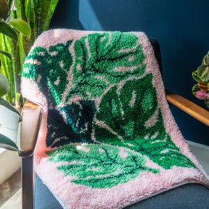 Könnte beinhalten: Ein rosa-grüner getufteter Textil mit einem großen Blattmuster. Die Blätter sind leuchtend grün mit weißen Akzenten, vor einem zarten rosa Hintergrund. Der Textil liegt über einem Stuhl und zeigt seine Textur und sein Design.