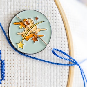 May include: A close-up of a needle and thread going through a white Aida cloth with a blue thread. A round enamel pin with a blue background and a yellow star, orange clouds, and a yellow shooting star is attached to the cloth.
