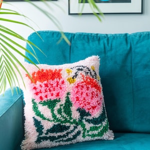 May include: A white throw pillow with a colorful floral design. The pillow features pink, red, orange, green, and yellow flowers on a white background. The pillow is sitting on a teal couch.