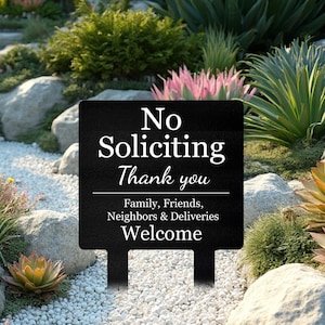 May include: A black rectangular metal garden sign featuring white text that reads: No Soliciting Thank you. Below a horizontal line, the text continues: Family, Friends, Neighbors & Deliveries Welcome. The sign has two stakes for ground insertion.