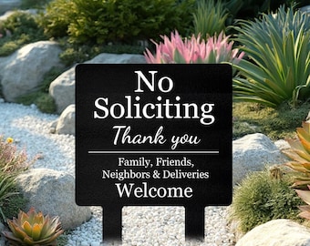 May include: A black rectangular sign with white text reading "No Soliciting Thank you". Below a line are the words "Family, Friends, Neighbors & Deliveries Welcome". The sign is set in a garden with rocks and plants.