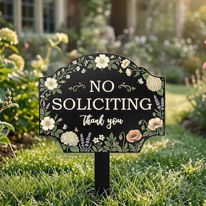 May include: A black garden sign with a scalloped top edge featuring white floral illustrations and the text NO SOLICITING Thank you in white lettering. The sign is mounted on a black stake and is positioned in a green grassy lawn.