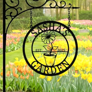May include: Black metal garden sign with the text "SASHA'S GARDEN" in a circular design, featuring a flower in a pot. The sign is suspended by chains from a decorative bracket. The sign also includes the text "EST 2025".