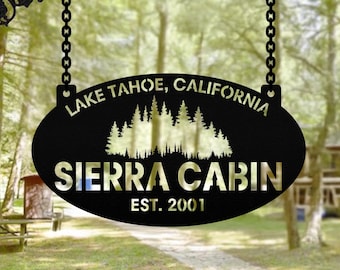 May include: Black metal sign with the text "LAKE TAHOE, CALIFORNIA" and "SIERRA CABIN EST. 2001". The sign features a forest silhouette and hangs from a decorative wrought iron bracket with chains.