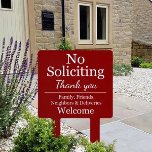 May include: A rectangular dark red garden sign with white text that reads No Soliciting Thank you, followed by Family, Friends, Neighbors & Deliveries Welcome. The sign features two stakes at the bottom for insertion into the ground.