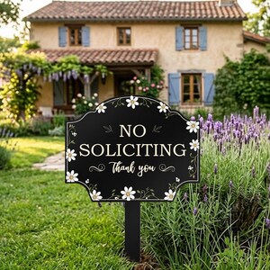 May include: A black yard sign with a white daisy border and the text "NO SOLICITING Thank you" in white lettering. The sign is mounted on a black stake and features a decorative, curved top edge with floral vine accents surrounding the text.