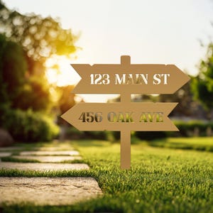 May include: A gold-colored metal signpost with two directional arrows. The top arrow reads "123 MAIN ST" and the bottom arrow reads "456 OAK AVE". The sign is placed in a grassy yard.