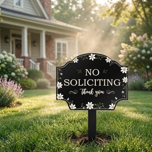 May include: A black yard sign with a scalloped edge featuring white daisy illustrations and green vines. The text reads NO SOLICITING in a bold serif font and Thank you in a cursive script. The sign is mounted on a black post.