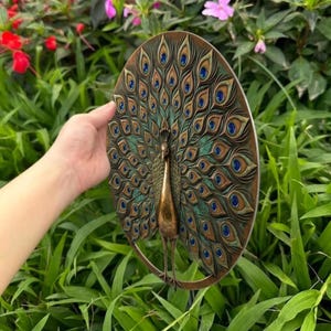 May include: A circular wall plaque featuring a three-dimensional bronze-toned peacock with its tail feathers fanned out. The feathers display intricate layered patterns in shades of bronze, copper, and teal, accented with deep blue circular centers.