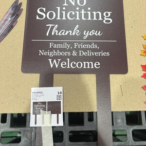 May include: A dark brown metal garden sign with white text that reads: No Soliciting Thank you, Family, Friends, Neighbours & Deliveries Welcome. The sign features two pointed stakes at the bottom for insertion into the ground.