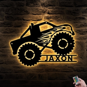 May include: Black metal monster truck wall art with the name "Jaxon" illuminated by warm yellow light. The truck features large, toothed wheels and flame details, mounted on a brick wall. A remote control is visible in the bottom right corner.