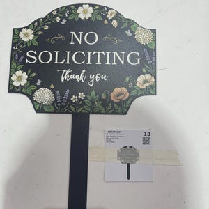 May include: A black yard sign with a pointed stake featuring the white text NO SOLICITING Thank you. The sign is decorated with a colorful floral border including white flowers, green leaves, and lavender sprigs. A small label with text is attached.