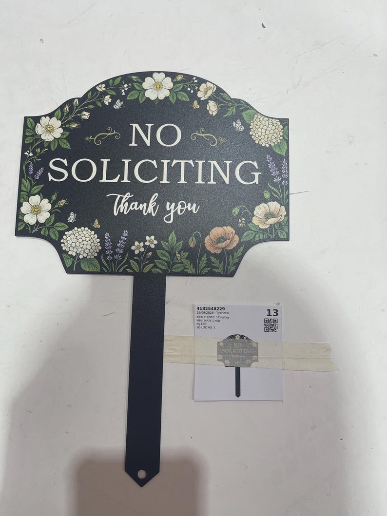 May include: A black yard sign with a pointed stake featuring the white text NO SOLICITING Thank you. The sign is decorated with a colorful floral border including white flowers, green leaves, and lavender sprigs. A small label with text is attached.