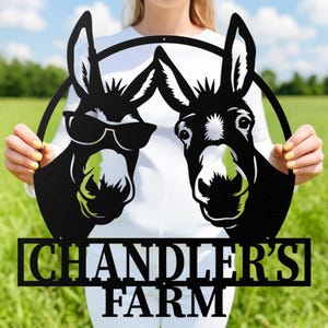 Puede incluir: Letrero de metal negro con dos caras de burro, una con gafas de sol. Los burros están dentro de un círculo, con las palabras "CHANDLER'S FARM" debajo. El letrero se sostiene frente a un campo verde.