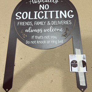 May include: A dark brown metal yard sign with an arched top and two stakes. The text reads: Absolutely NO SOLICITING, FRIENDS, FAMILY & DELIVERIES always welcome, If that's not YOU, Do not knock or ring bell. A small product label is attached.