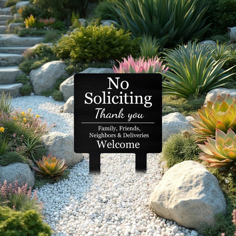 May include: A black rectangular metal garden sign featuring white text that reads: No Soliciting Thank you. Below a horizontal line, the text continues: Family, Friends, Neighbors & Deliveries Welcome. The sign has two stakes for ground insertion.