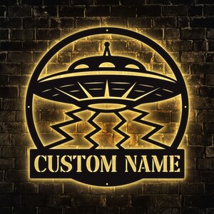 May include: Black metal UFO sign with the text "CUSTOM NAME" on a rectangular banner. The sign features a detailed depiction of a flying saucer with a glowing yellow light, set against a dark brick wall.