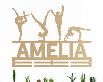 May include: Gold metal gymnastics medal holder with the name Amelia and four gymnast silhouettes. The holder has a bar for hanging medals and a row of hooks below.