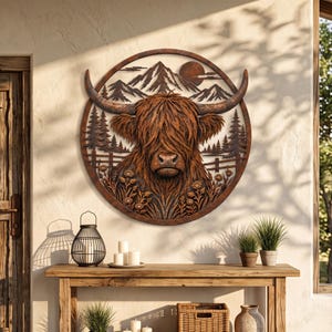 May include: A circular metal wall art piece featuring a detailed Highland cow head in the center. The design includes a mountain range, a sun, pine trees, a fence, and wildflowers. The artwork has a rustic, weathered brown finish.