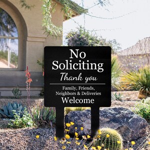 May include: A black rectangular metal garden sign with white text that reads: No Soliciting Thank you. Family, Friends, Neighbors & Deliveries Welcome. The sign features two pointed stakes at the bottom for insertion into the ground.