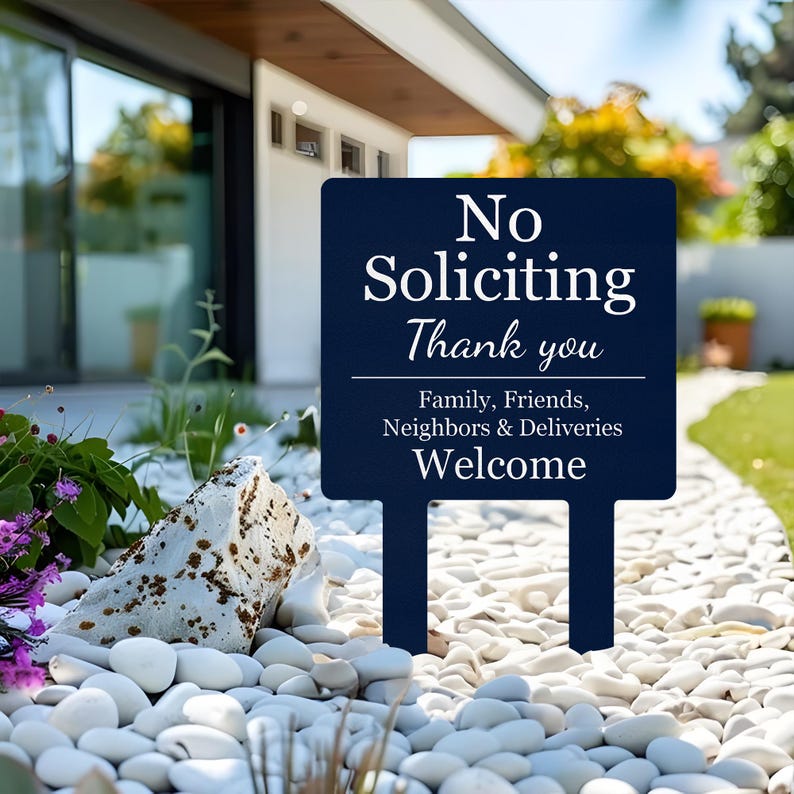 May include: A dark blue rectangular garden sign featuring white text that reads: No Soliciting Thank you. Below a horizontal line, the text continues: Family, Friends, Neighbors & Deliveries Welcome. The sign has two stakes for ground insertion.