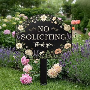 May include: A black garden sign with a scalloped top edge featuring cream, peach, and green floral illustrations. The text reads NO SOLICITING in large capital letters and Thank you in a smaller script font, mounted on a black stake.