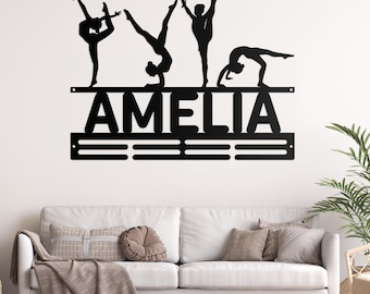 May include: Black metal gymnastics medal hanger with the name Amelia and five gymnast silhouettes. The hanger has a bar for hanging medals and a row of hooks below.