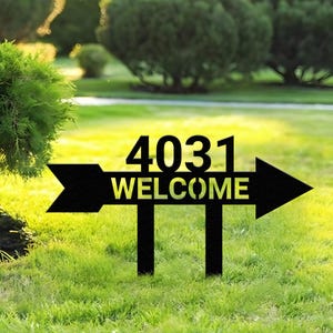 Puede incluir: Señal de jardín de metal negro con el número "4031" y la palabra "WELCOME" en un diseño de flecha. La señal está colocada en un jardín de césped, con árboles al fondo.