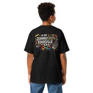 Op de afbeelding: Zwart T-shirt met een kleurrijk grafisch ontwerp op de achterkant. Het ontwerp bevat de tekst "IN MY JOHNNY KNOXVILLE ERA" met verschillende illustraties zoals een skateboard, hart en oneindigheidssymbool. Het shirt is gemaakt van een zacht materiaal.