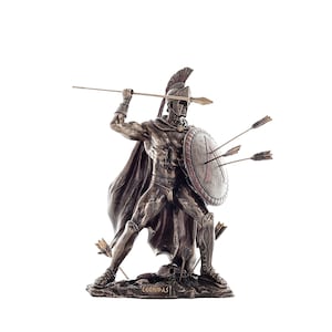 Può includere: Statua in bronzo di un guerriero spartano in posizione di combattimento, con lancia e scudo. Lo scudo presenta impatti di frecce. Il guerriero indossa elmo e mantello. La base della statua riporta il nome "LEONIDAS".