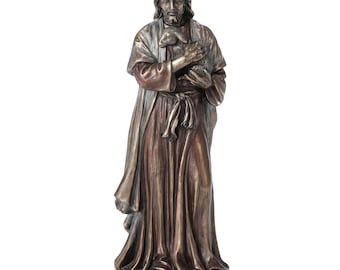 Puede incluir: Estatua de color bronce de una figura con pelo largo y barba, vestida con túnicas fluidas y sosteniendo un cordero. La estatua tiene un diseño detallado y se apoya sobre una base redonda. La expresión de la figura es serena.