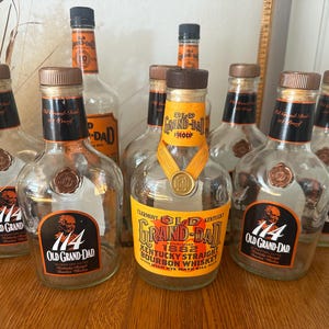 Puede incluir: Una colección de botellas vacías de whisky bourbon Old Grand-Dad. Las botellas son de vidrio transparente con etiquetas negras y naranjas. Las etiquetas presentan el texto "114 Old Grand-Dad" y "Kentucky Straight Bourbon Whiskey". Las botellas tienen tapas marrones.
