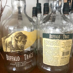 May include: Clear glass Buffalo Trace Kentucky Straight Bourbon Whiskey bottles with green caps. The labels feature a buffalo illustration and text. Several bottles are visible, some in focus, others blurred in the background.