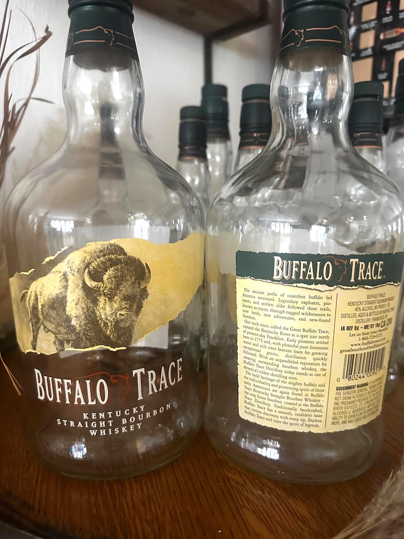 May include: Clear glass Buffalo Trace Kentucky Straight Bourbon Whiskey bottles with green caps. The labels feature a buffalo illustration and text. Several bottles are visible, some in focus, others blurred in the background.