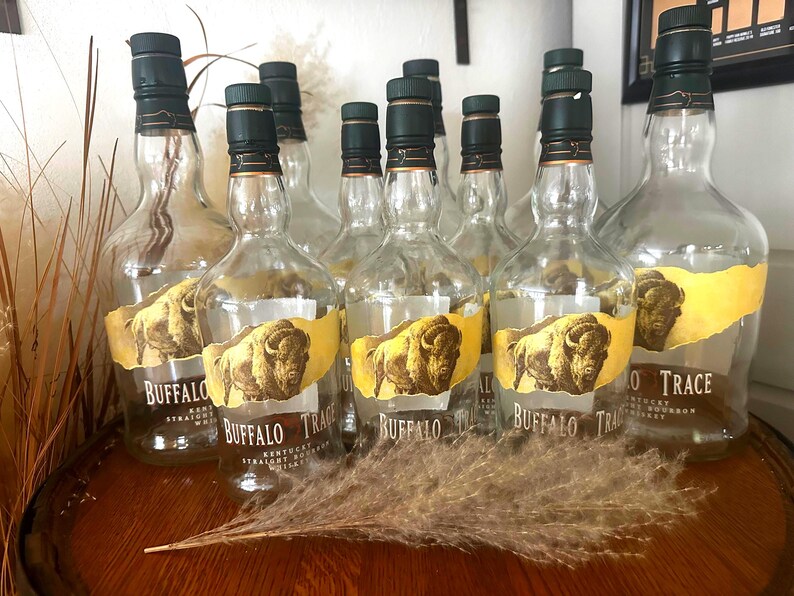 May include: A collection of empty Buffalo Trace bourbon bottles with green caps and yellow labels featuring a bison illustration. The bottles are arranged on a wooden surface, with dried ornamental grass in the foreground.