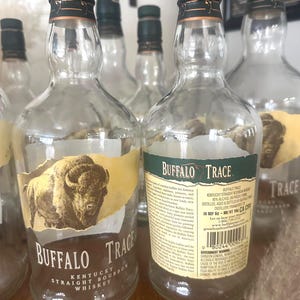 May include: Several empty Buffalo Trace Kentucky Straight Bourbon Whiskey bottles. The clear glass bottles have green and gold labels with the brand name and a bison illustration. The bottles are arranged on a wooden surface.